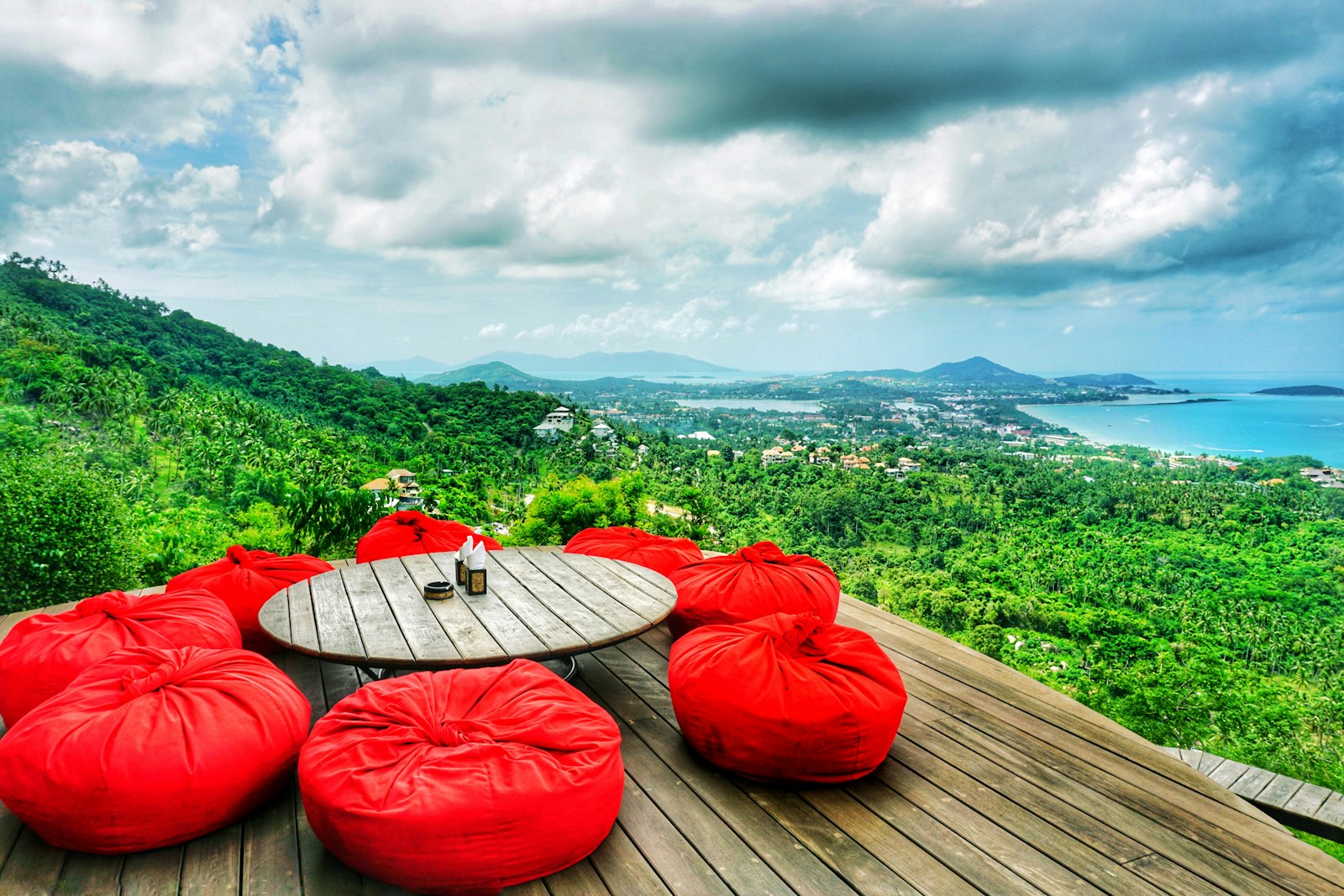A round wooden table with red floor cushions on a deck above the jungle, looking over Chaweng and the east coast of Koh Samui