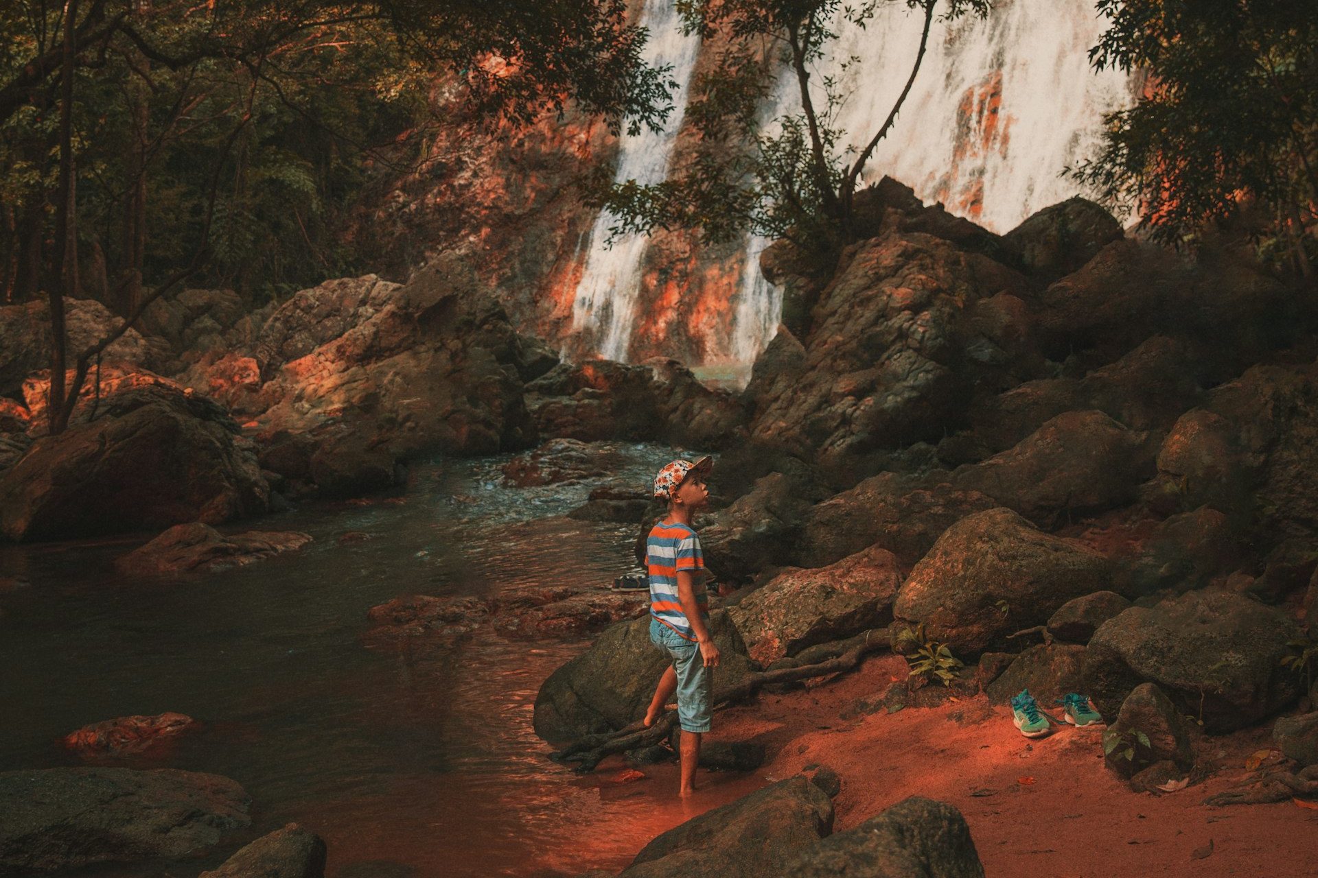 A child in a striped top standing on the rocks below a tall jungle waterfall, with the falls lit orange by the afternoon light